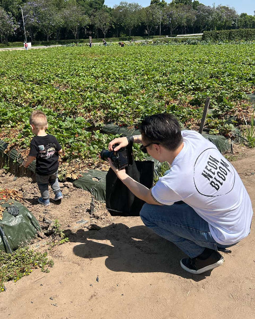 Man and child in a strawberry field with a camera, wearing 'Full Bloom' t-shirts.
