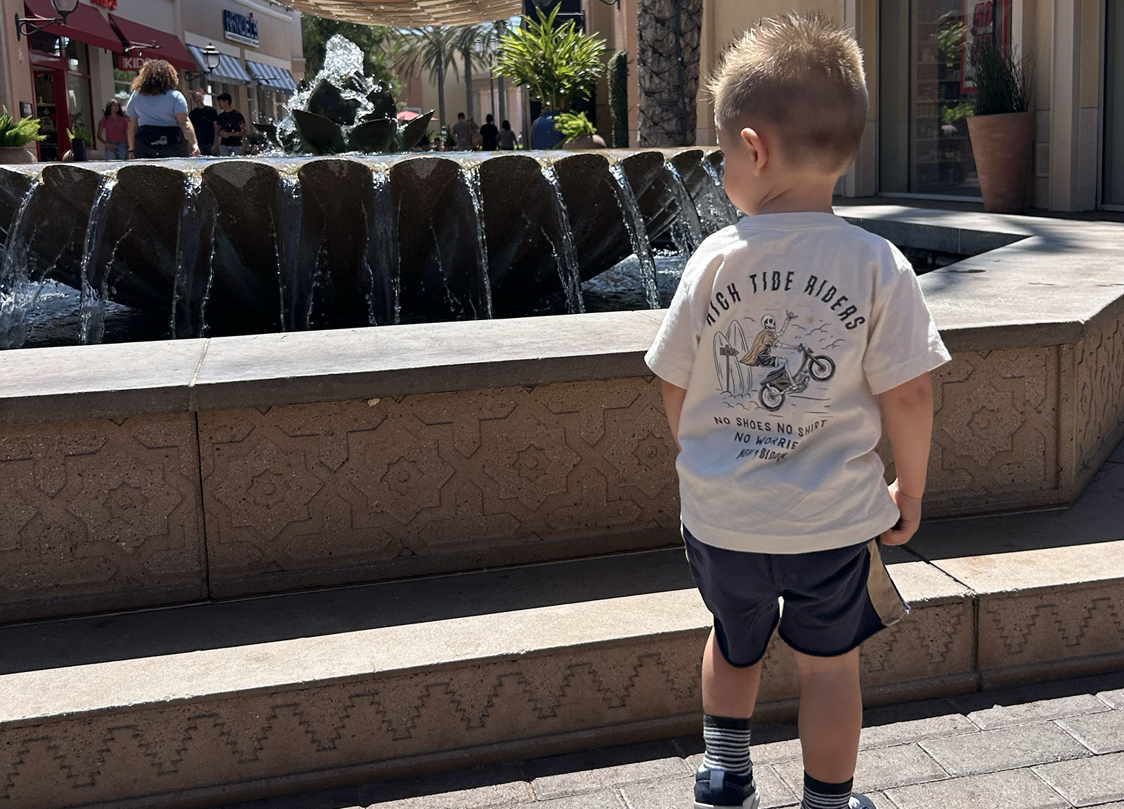 Child standing in front of a water fountain with a 'High Tide Riders' t-shirt.