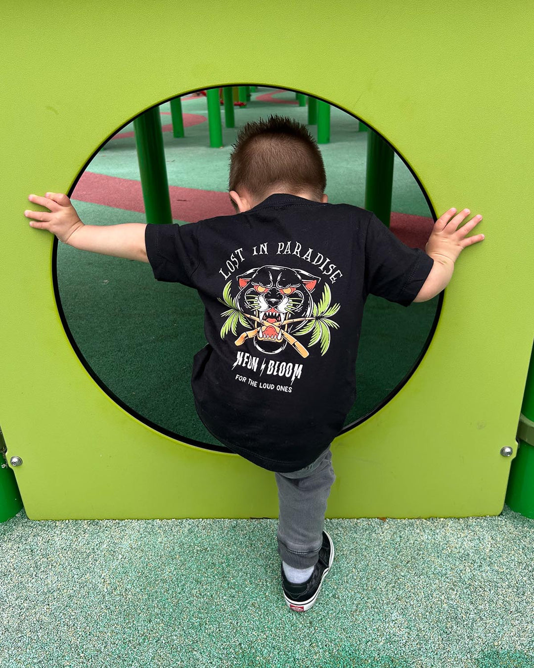 Child wearing a black t-shirt with a graphic design, standing in front of a green playground structure.